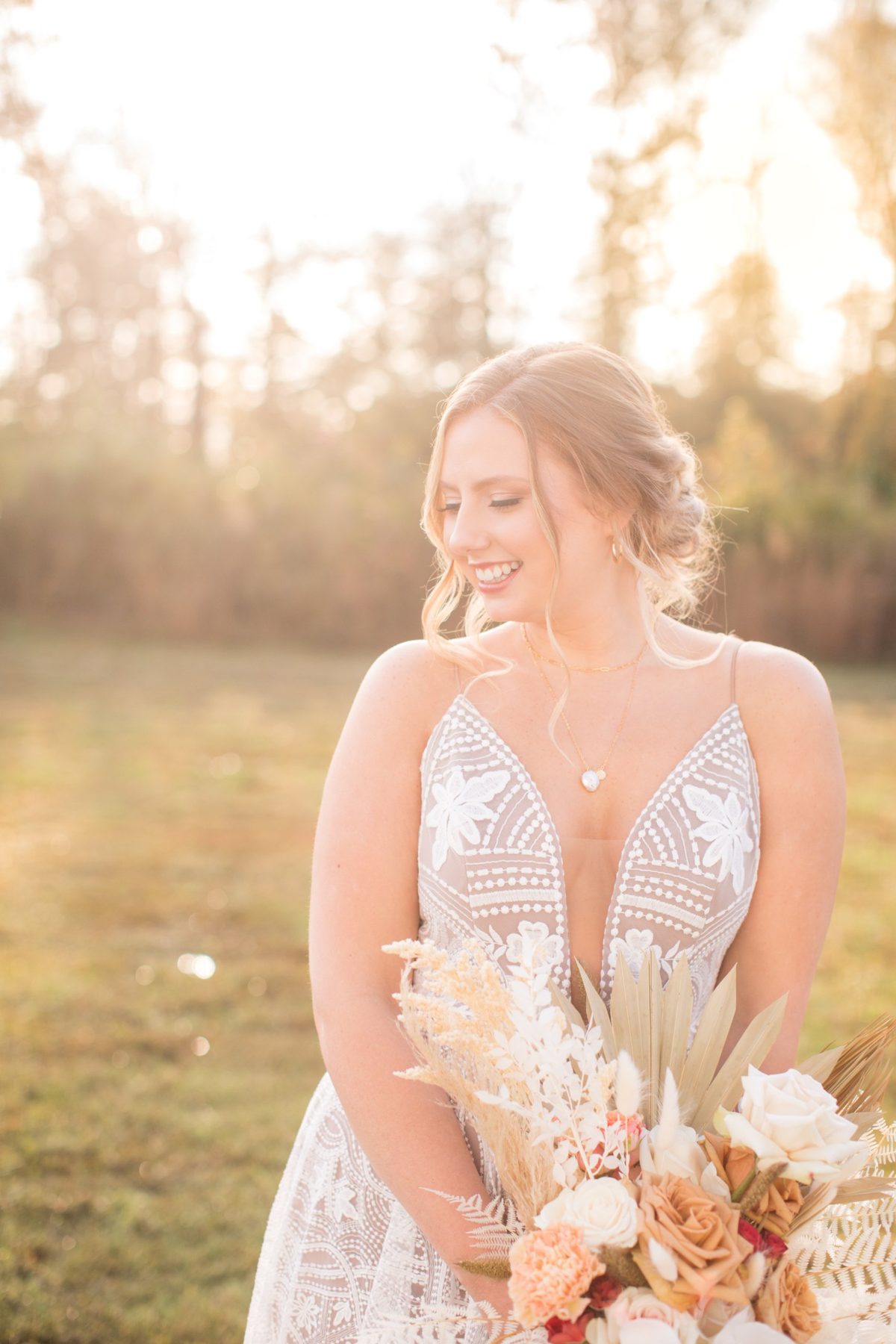 A bride smiling in a lace gown with a dried-flower bouquet, lit by golden-hour sun on the estate lawn