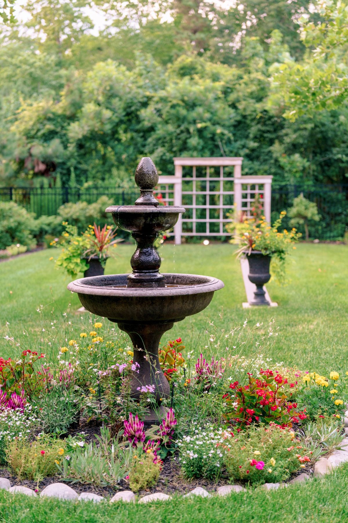 Stella's Secret Garden: a tiered stone fountain ringed with flowers, a white trellis and iron fence behind it