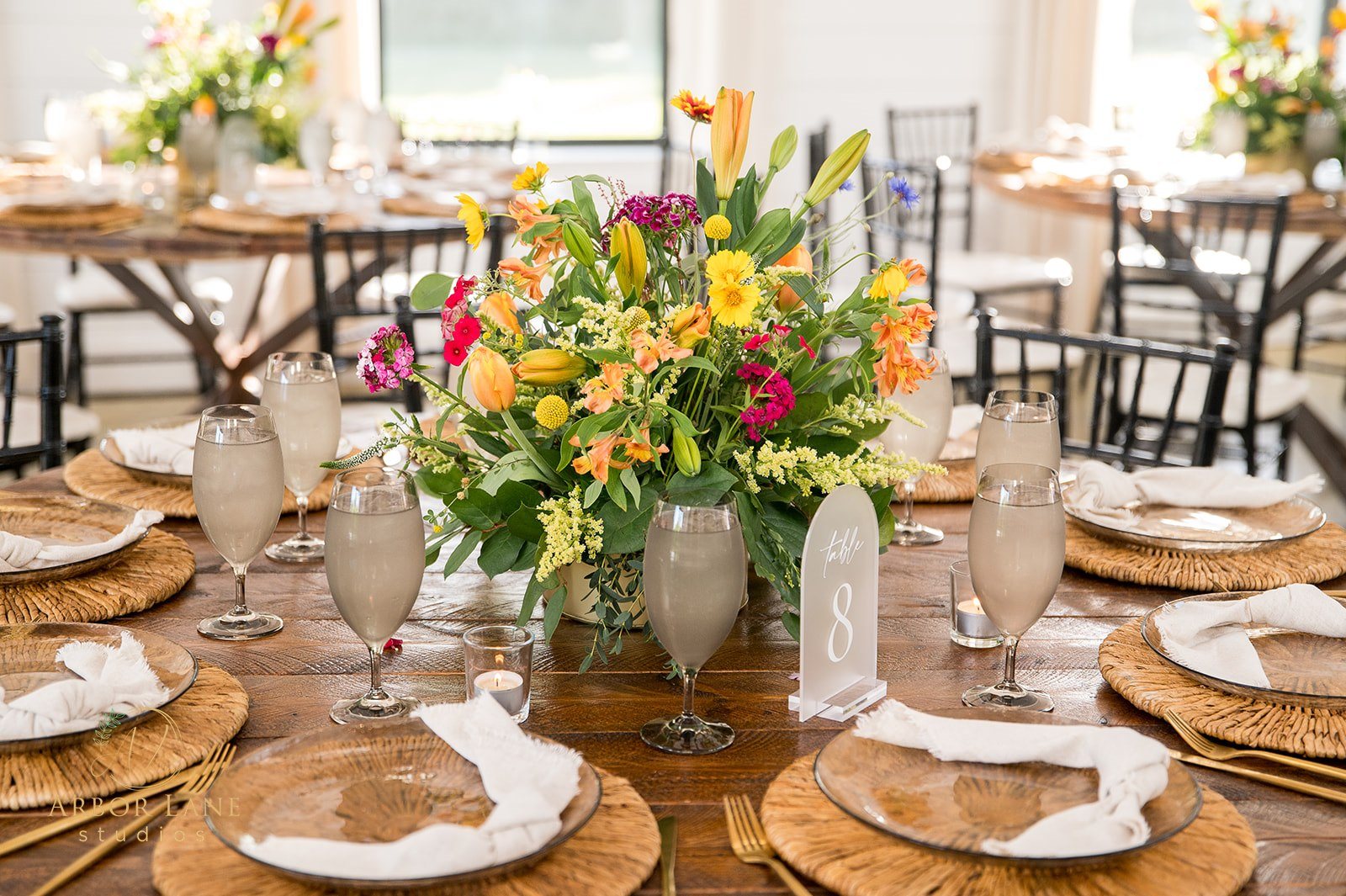 A reception table set with a bright wildflower centerpiece, woven chargers, glass plates, linen napkins and gold flatware